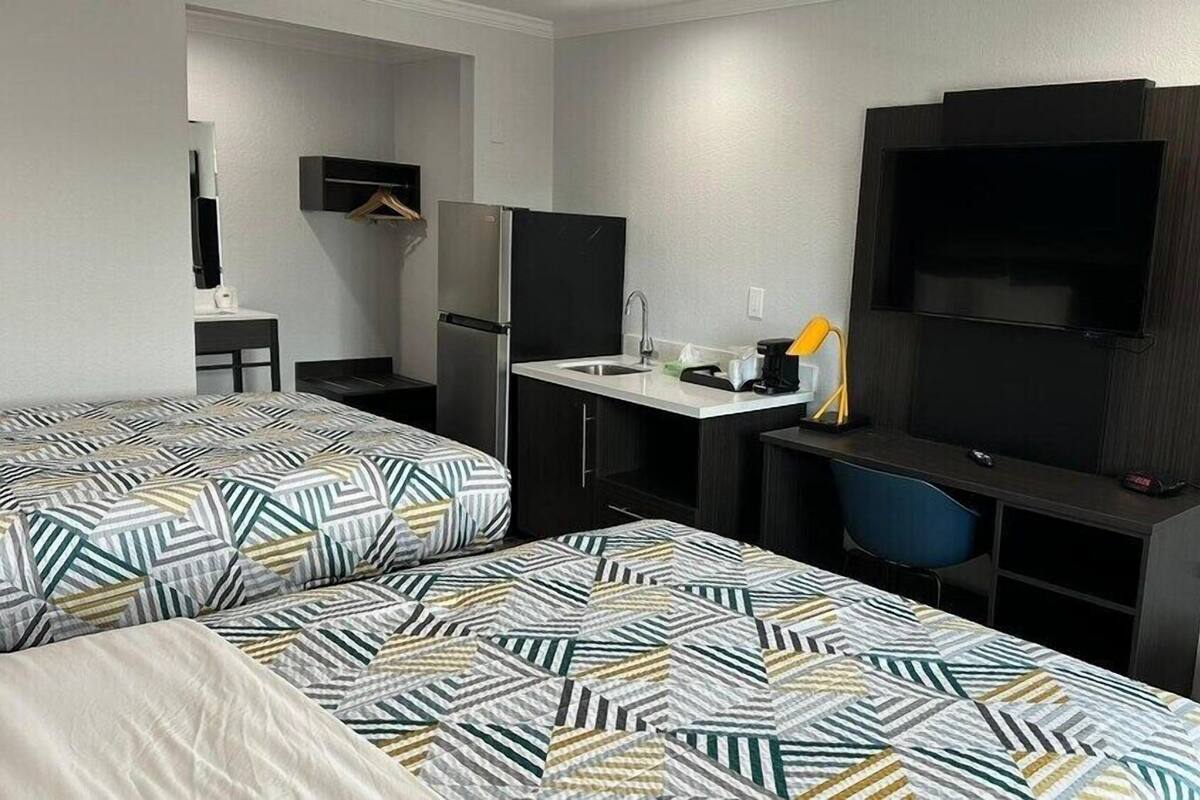 A hotel room with two beds, a desk with a chair, a TV, and a small kitchenette.
