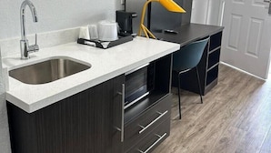 Eine kompakte Küchenzeile mit Spüle aus Edelstahl, integriertem Backofen, kleiner Arbeitsfläche und einem schwarzen Stuhl.