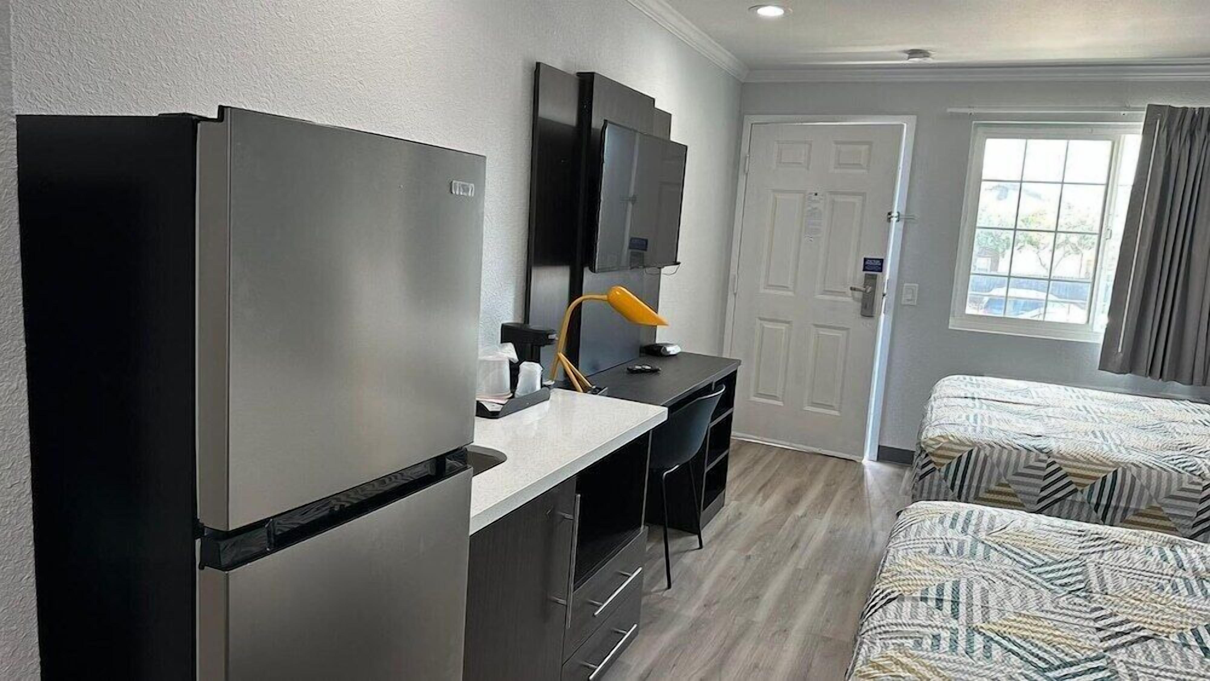 A hotel room with a kitchenette, a desk with a chair, a bed with patterned bedding, a television, and a window with curtains.