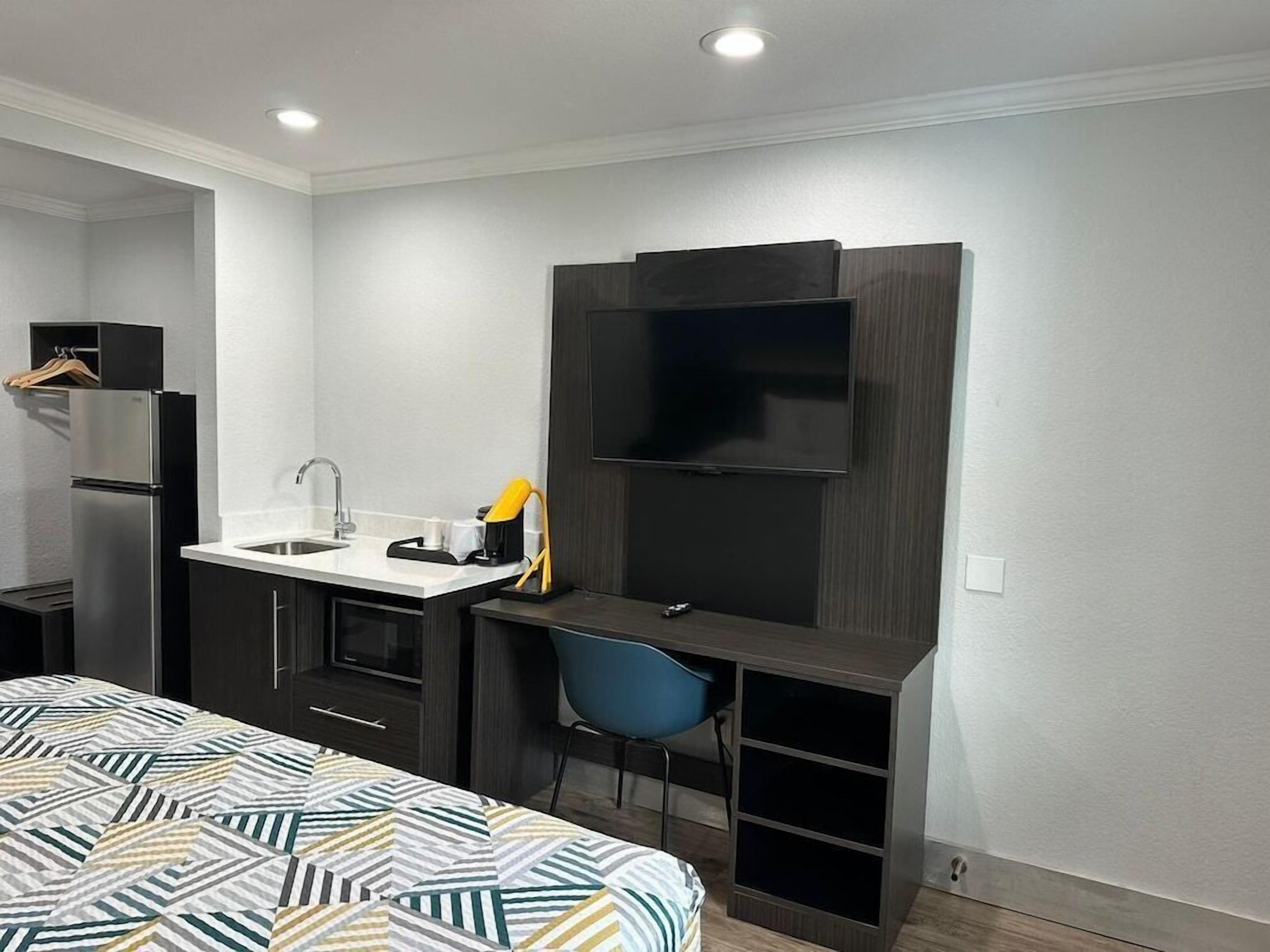 Habitación de hotel pequeña con cama, kitchenette, escritorio con silla, televisor montado en la pared y un estante.