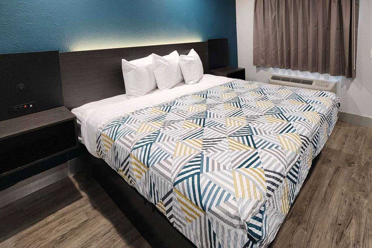 A hotel room with a bed, a nightstand, a wall-mounted air conditioner, a door, and a window with curtains.