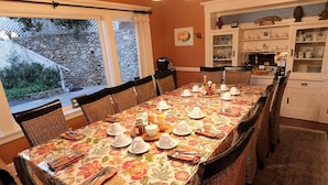Un comedor con una mesa larga dispuesta para una comida, rodeada de sillas de mimbre, y una cocina al fondo.