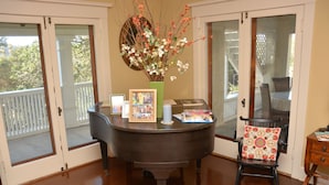 Un comedor con un piano, una silla con cojín floral, un jarrón con flores y un cuadro en el piano.