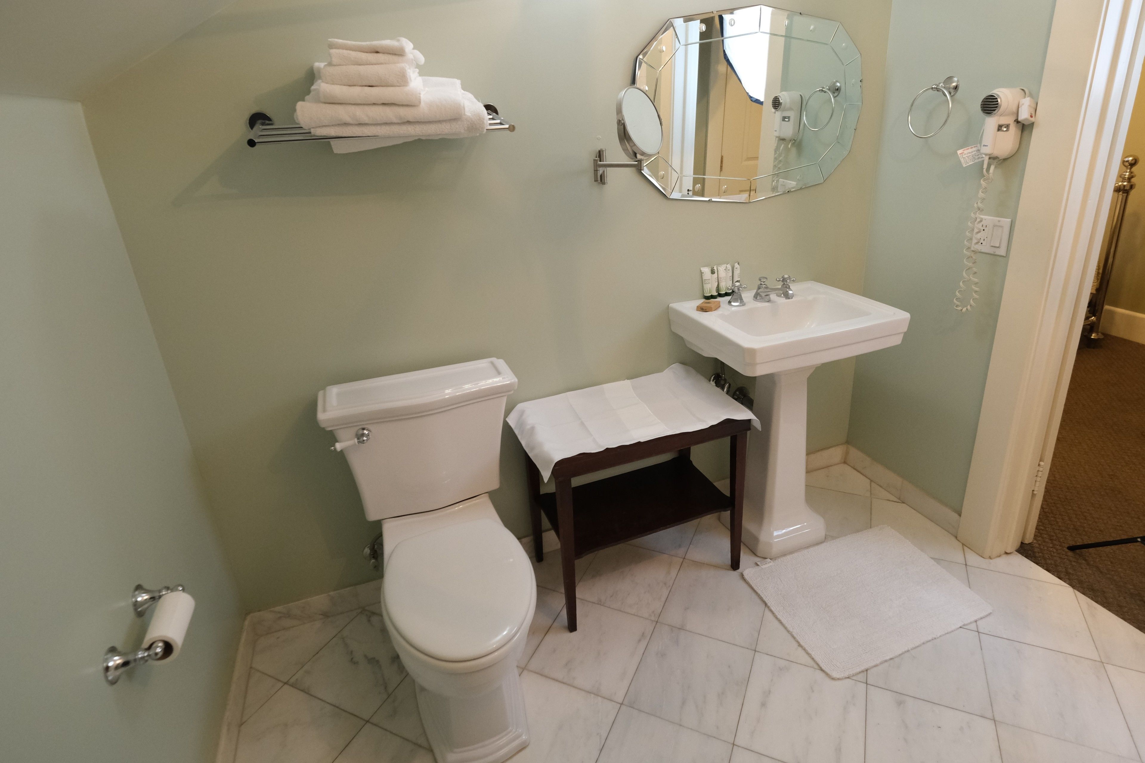 Ein Badezimmer mit Toilette, Waschbecken und Spiegel. Handtücher befinden sich an einem Ablagegestell, ein Föhn und ein Telefon sind an der Wand angebracht.