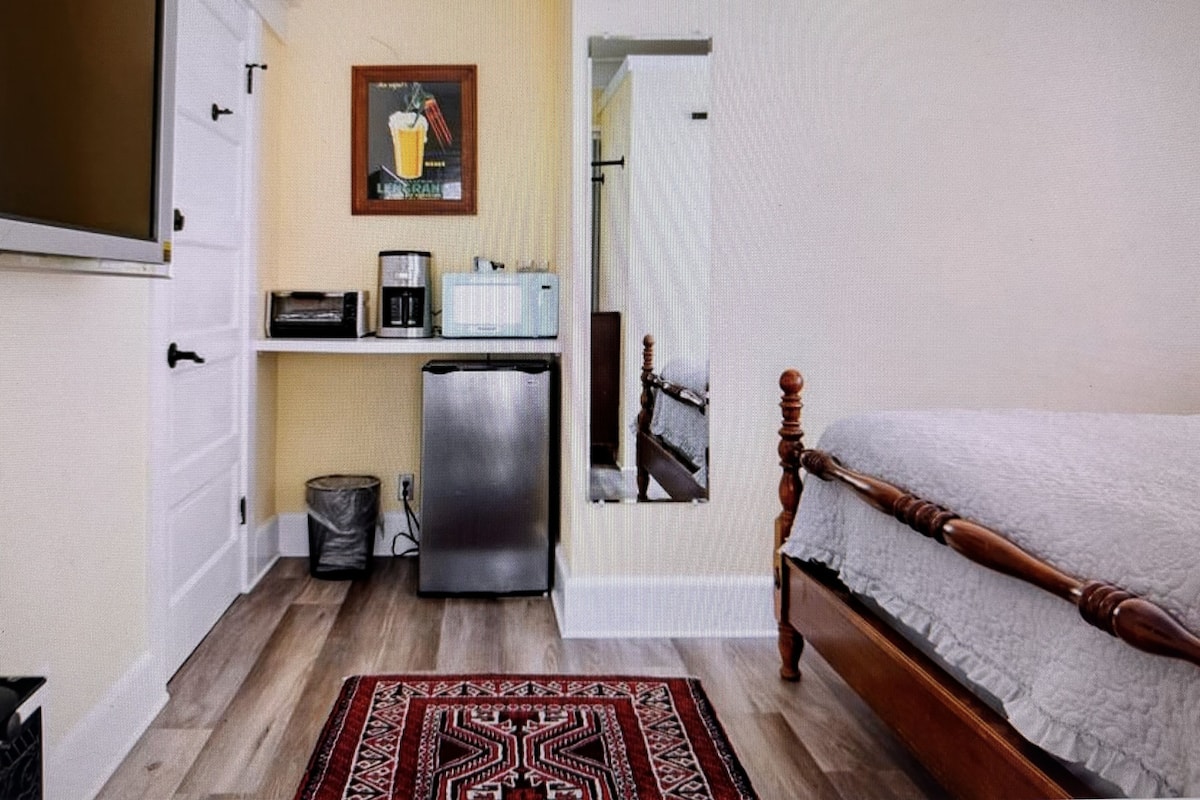 Ein kleines Zimmer mit einem Bett, einer kompakten Küchenzeile inklusive Kühlschrank, Mikrowelle und Kaffeemaschine sowie einem Teppich mit Muster.