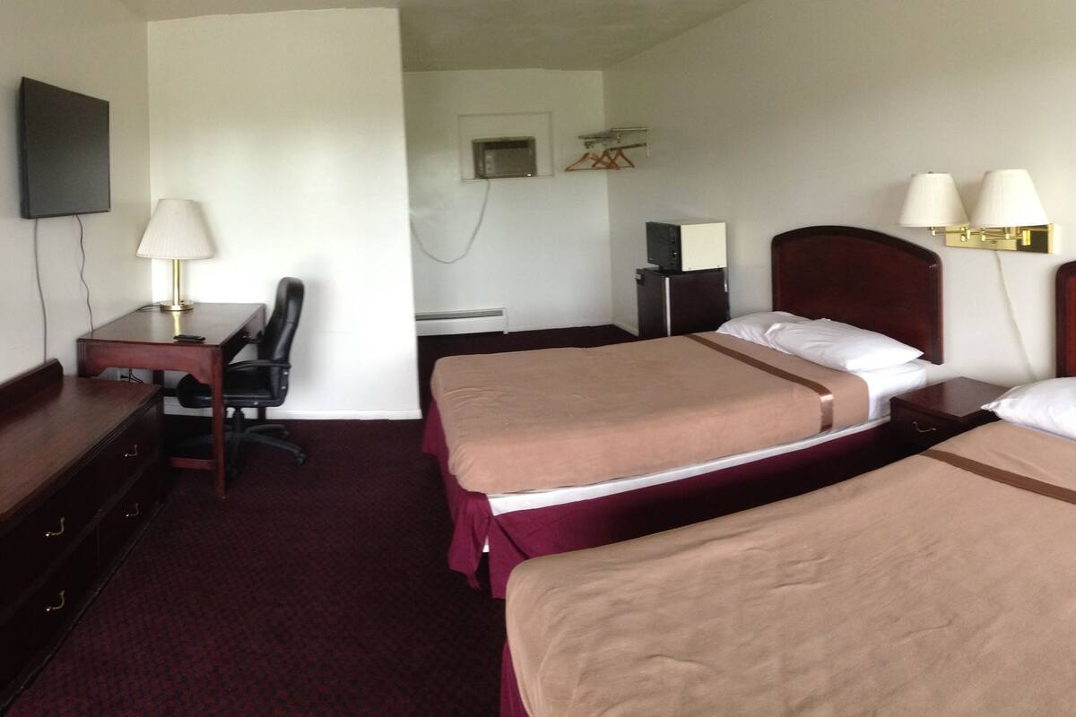 A hotel room with two beds, a desk, a chair, a TV, and a lamp.