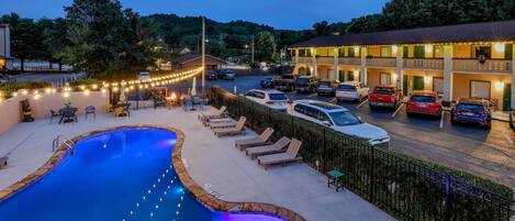 A swimming pool with lounge chairs and a covered seating area, surrounded by a parking lot with cars.