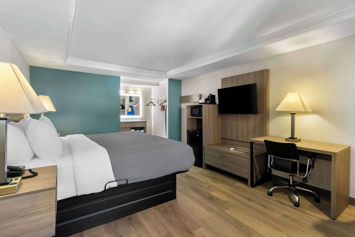 A modern hotel room with a bed, desk, chair, TV, and a small kitchenette.