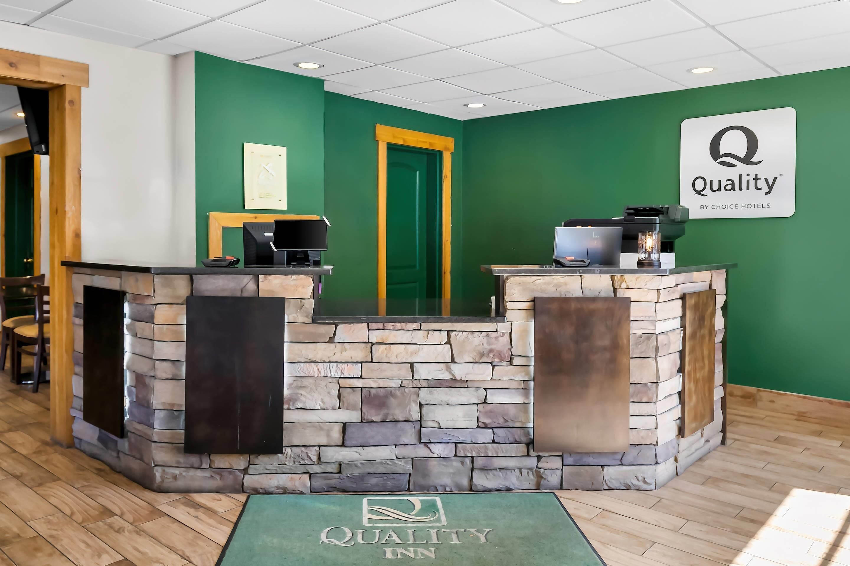 Zona de recepción con mostrador de piedra, una mesa de recepción y una pared verde con el letrero 'Quality Inn'.