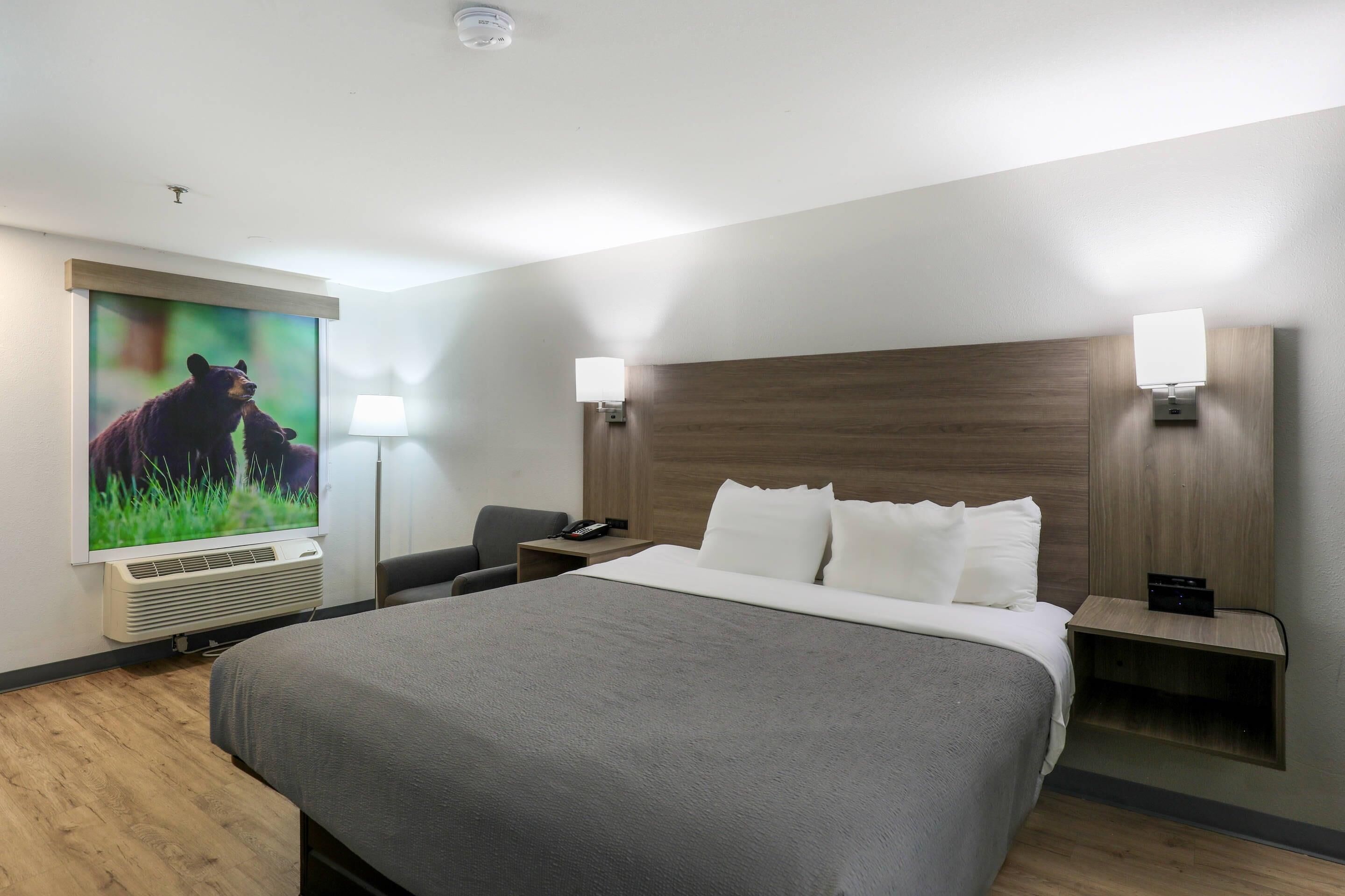 A hotel room with a large bed, a desk, a chair, a telephone, a lamp, and a wall-mounted air conditioner.