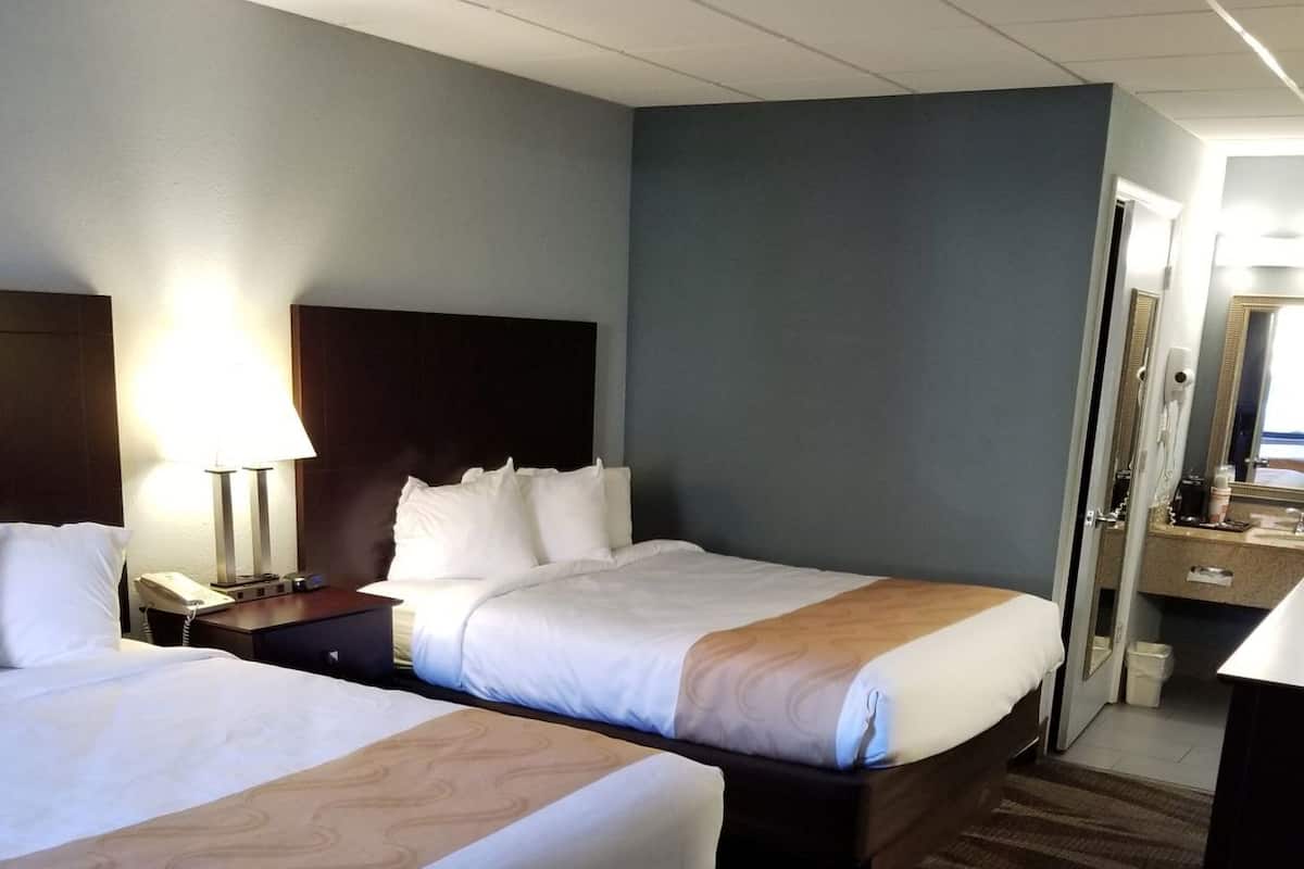 A hotel room with two beds, a nightstand with a lamp, a desk with a phone, and a bathroom visible through an open door.