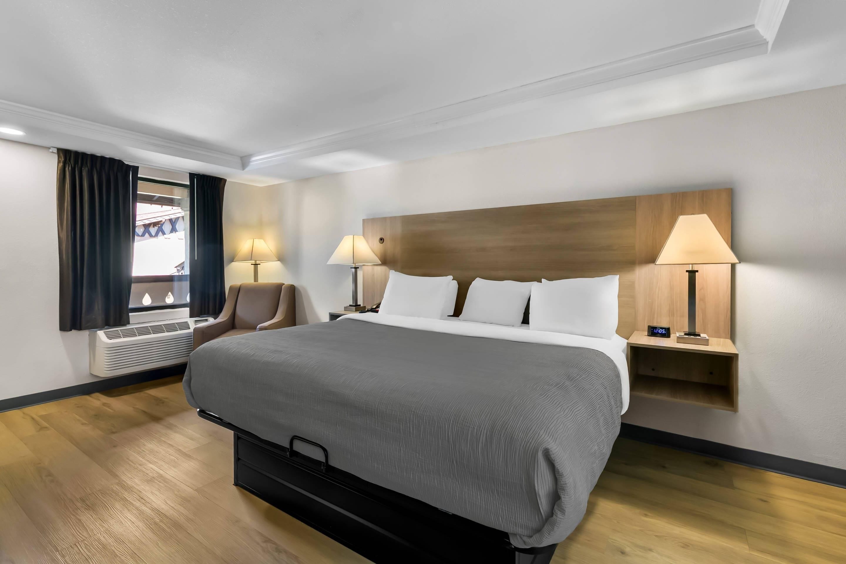 A hotel room with a large bed, two bedside tables with lamps, a chair, a window with curtains, and an air conditioning unit.