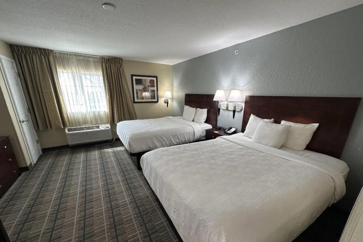 A hotel room with two beds, a carpeted floor, a window with curtains, and wall-mounted lamps.