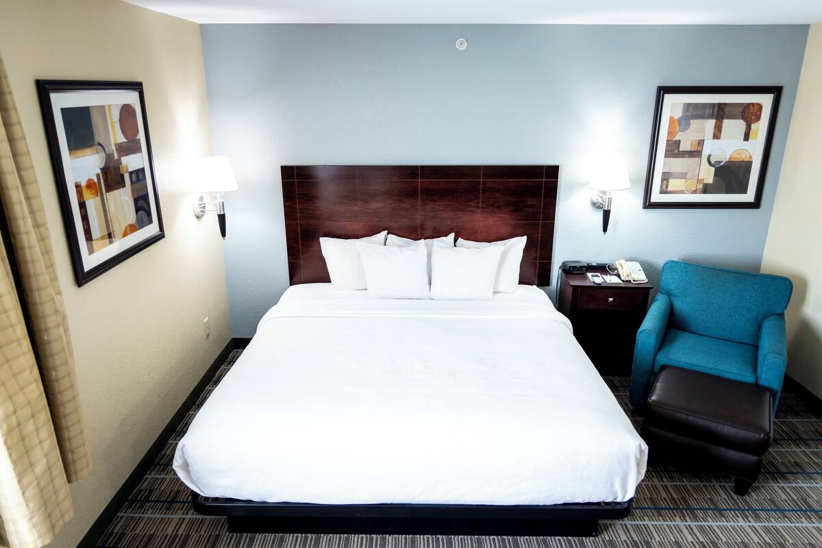 Une chambre d’hôtel avec un grand lit, deux tables de chevet, un fauteuil bleu et un pouf marron.