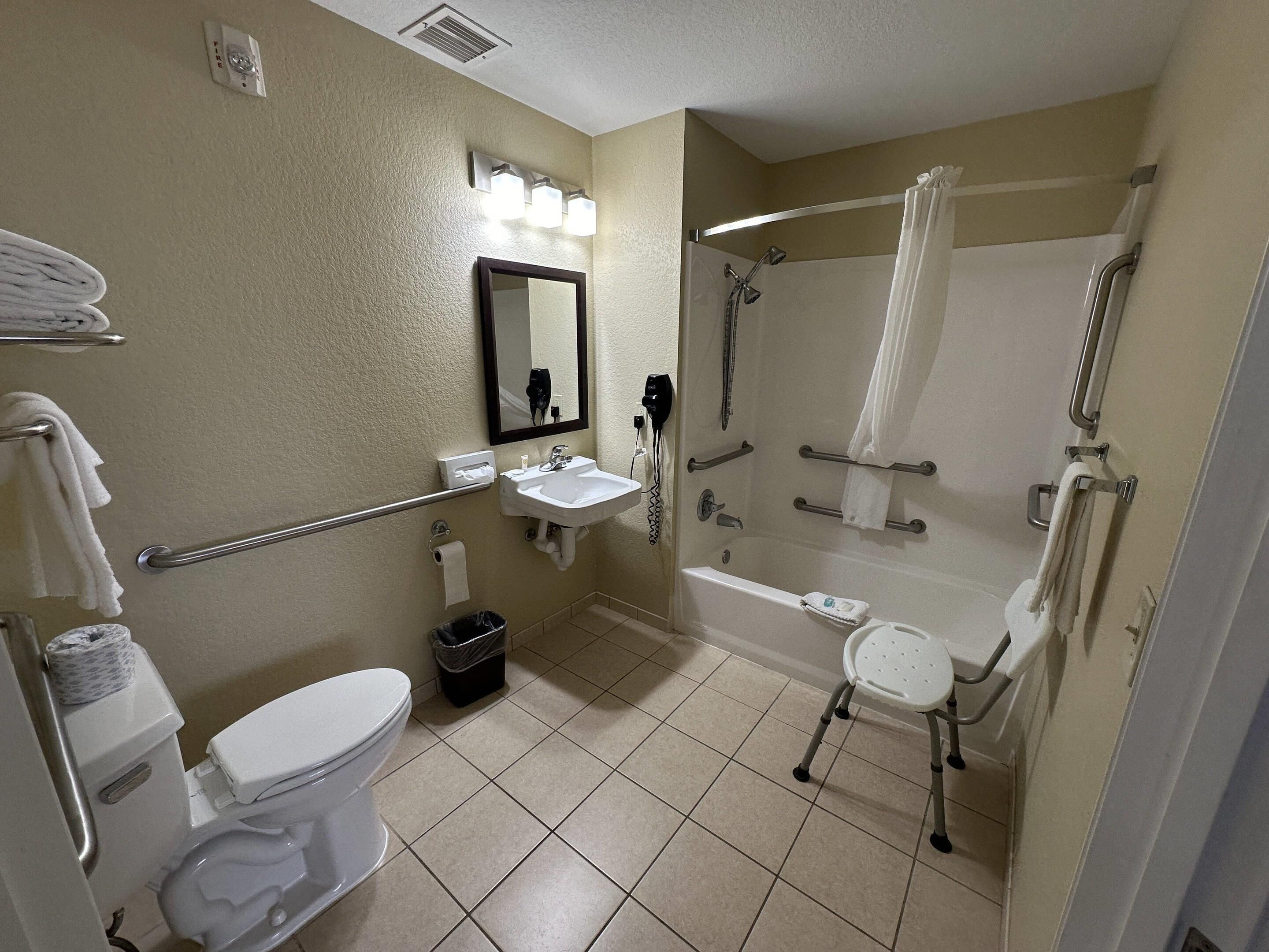 Un baño de hotel estándar con inodoro, lavamanos, espejo, ducha y barras de apoyo.