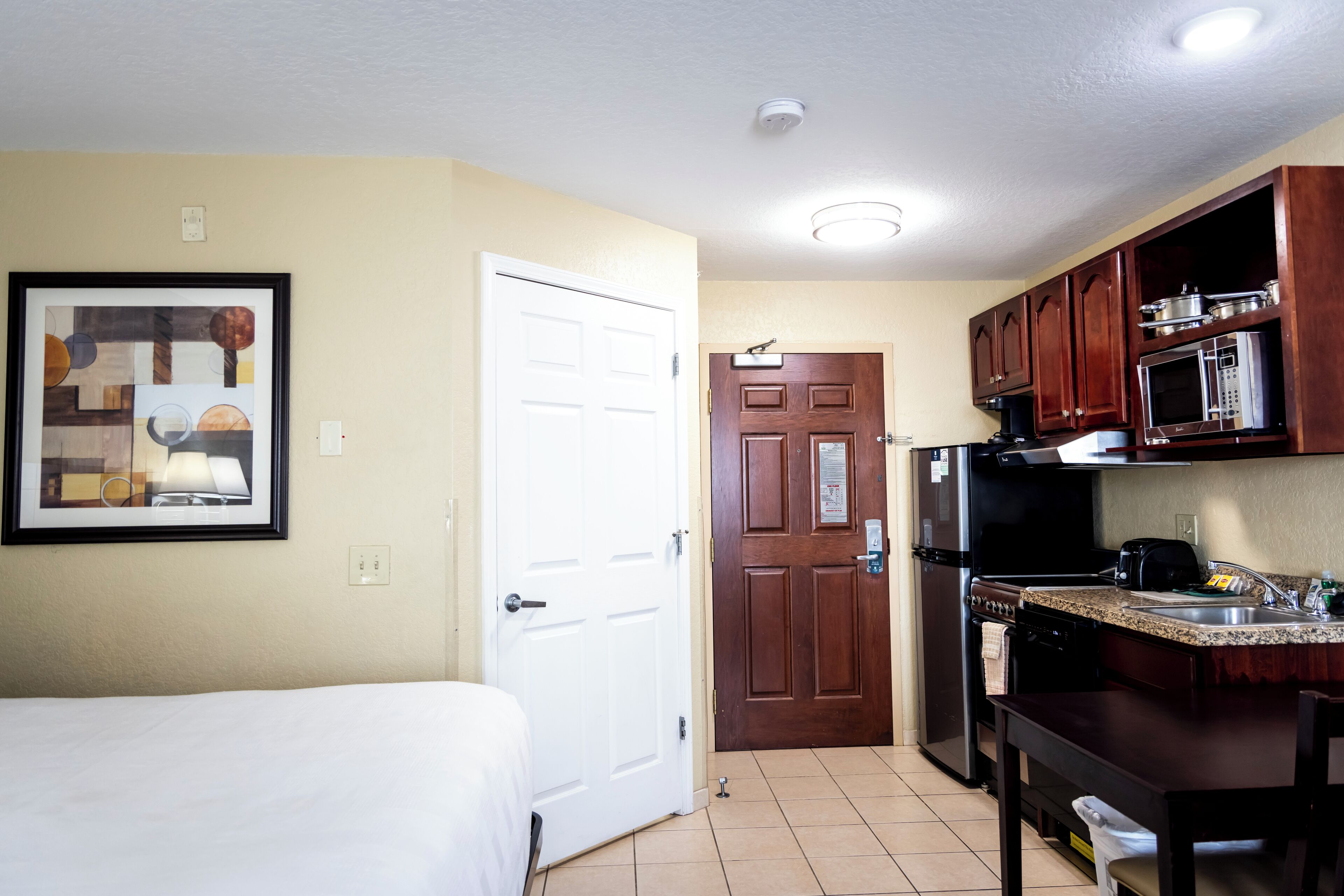 A compact hotel room with a bed, a kitchenette, and a door leading to another room.