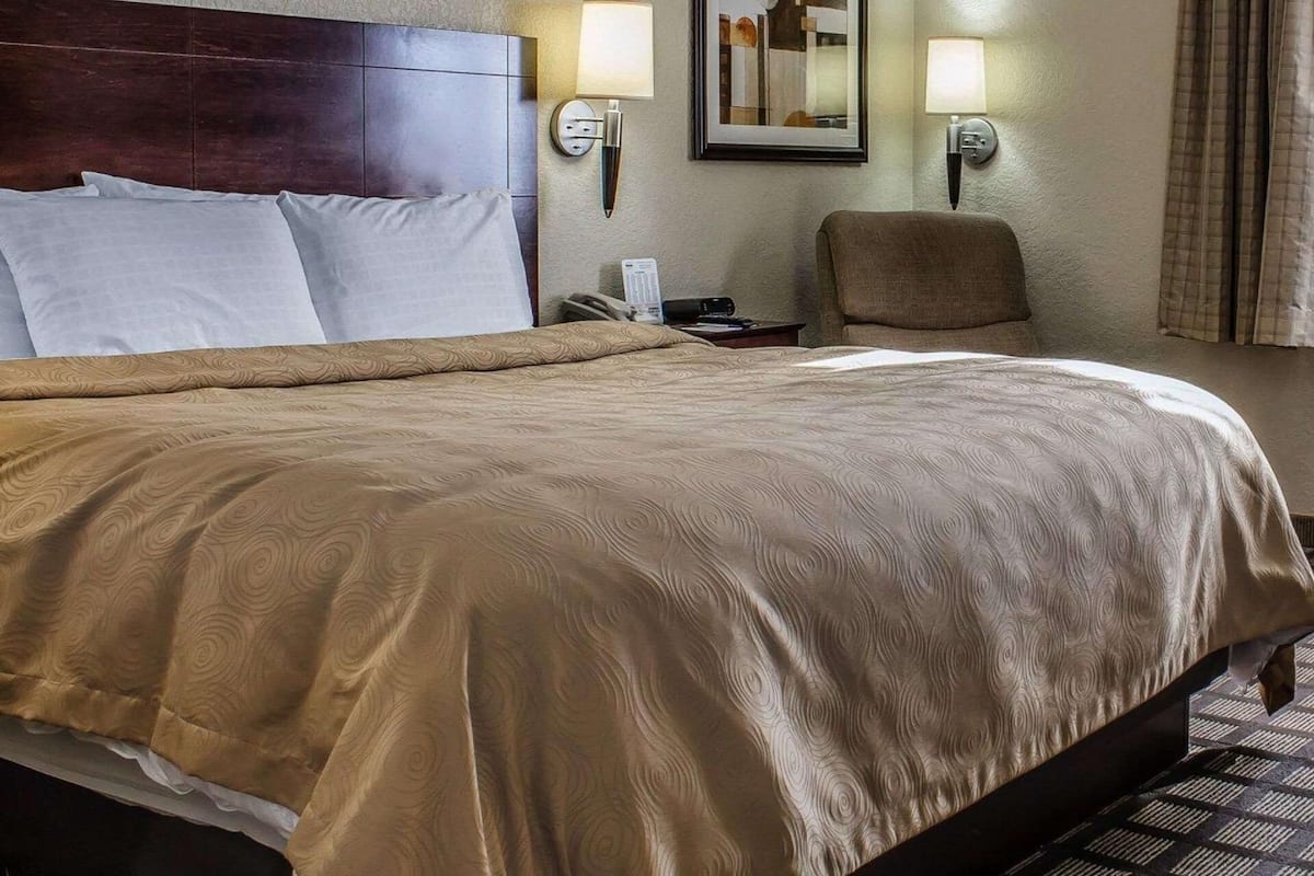 Une chambre d’hôtel avec un grand lit, deux lampes fixées au mur, un tableau encadré, une chaise et un téléphone.
