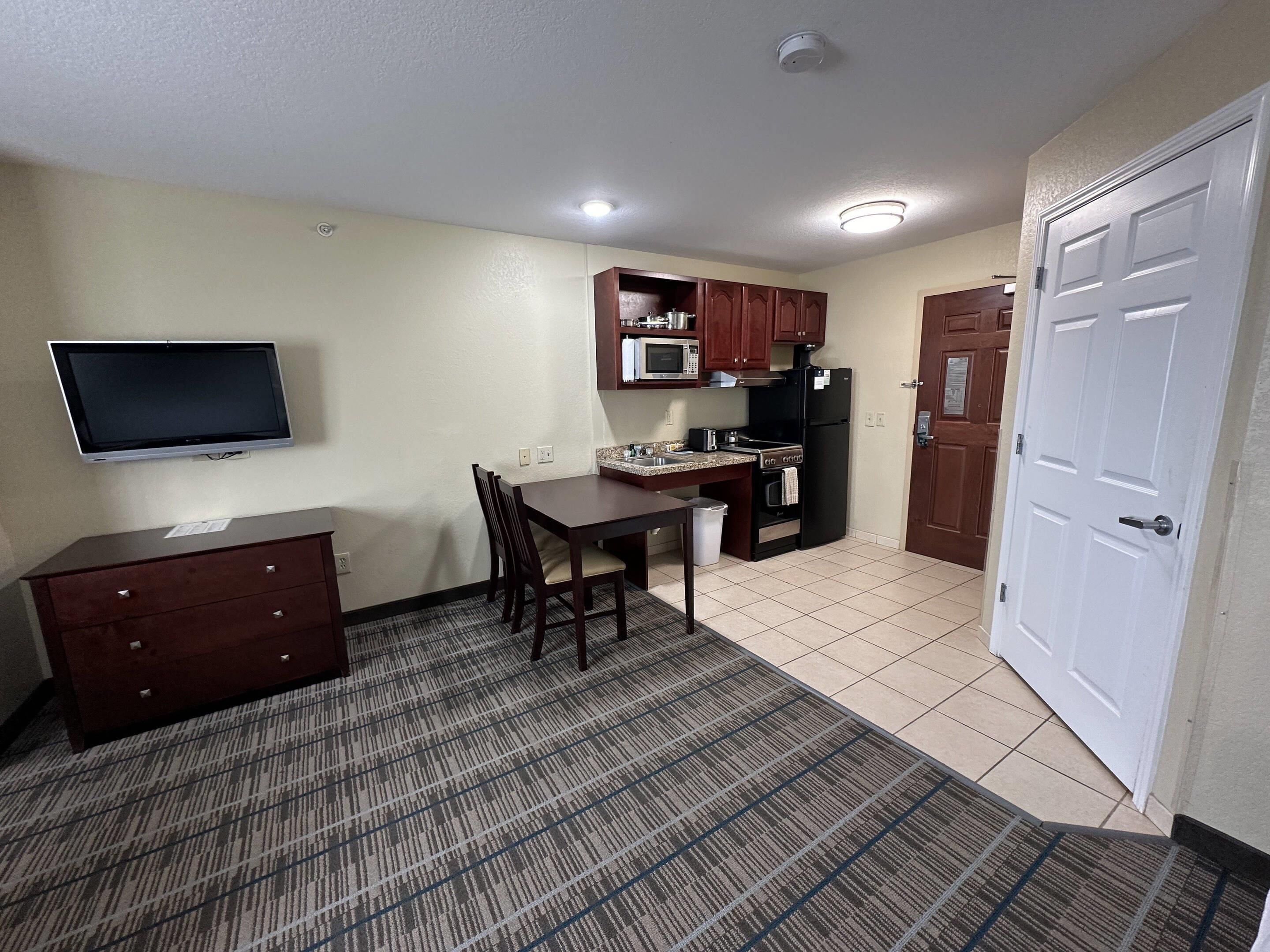 A compact kitchen with a microwave, stove, and refrigerator, a dining table with chairs, a TV mounted on the wall, and a door with a handle.