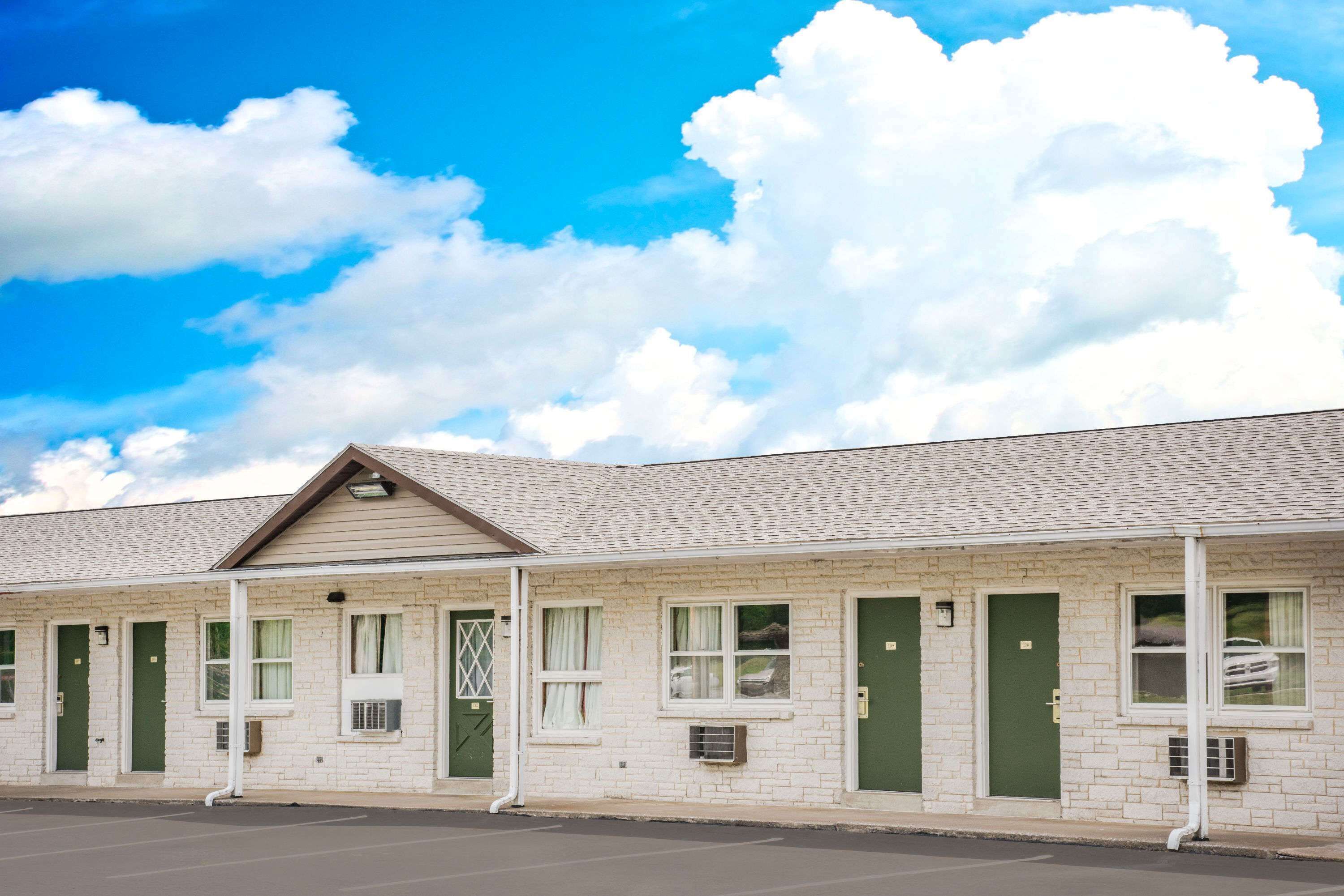 A row of motel rooms with individual doors, a parking lot, and a clear blue sky with clouds.