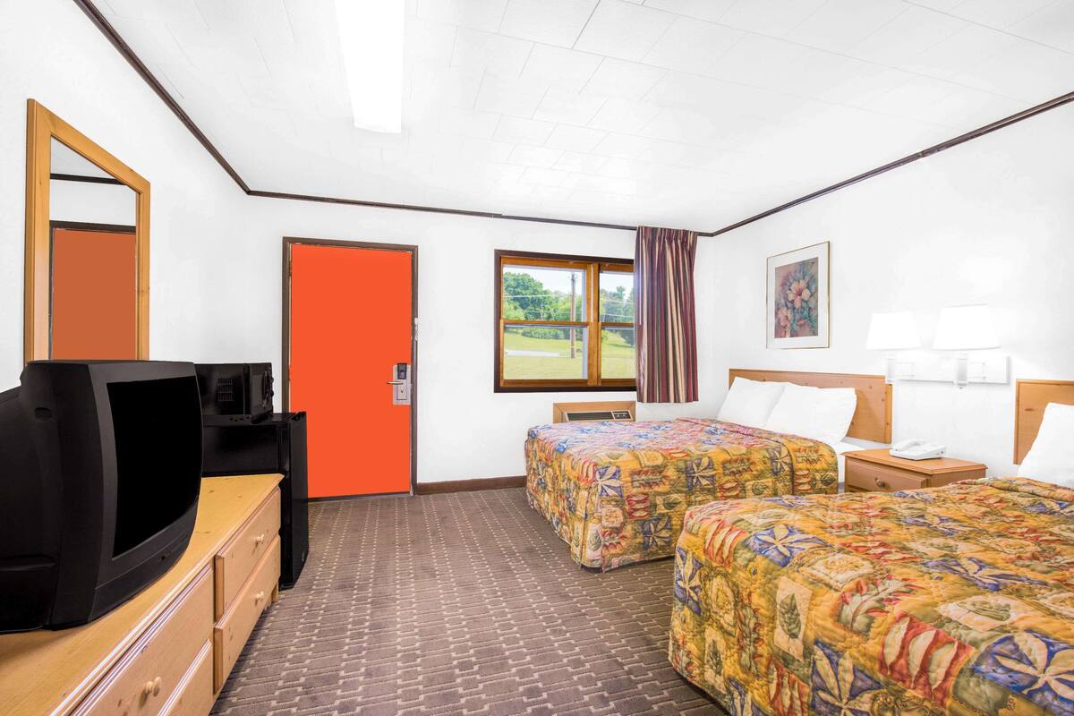 Ein Hotelzimmer mit zwei Betten, einem Fernseher, einem Schreibtisch mit Schubladen, einer orangefarbenen Tür, einem Fenster mit Blick und einem Bild an der Wand.