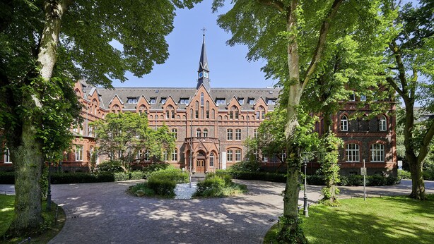 A historic building with a prominent tower and arched windows, surrounded by a cobblestone pathway and lush greenery. - Hotel MutterHaus Düsseldorf (Düsseldorf)