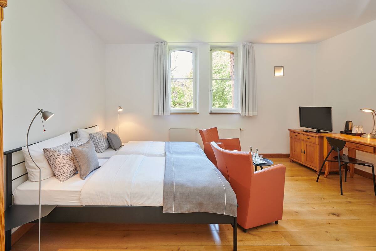 A bedroom with a bed, a desk, a chair, a TV, and two windows.