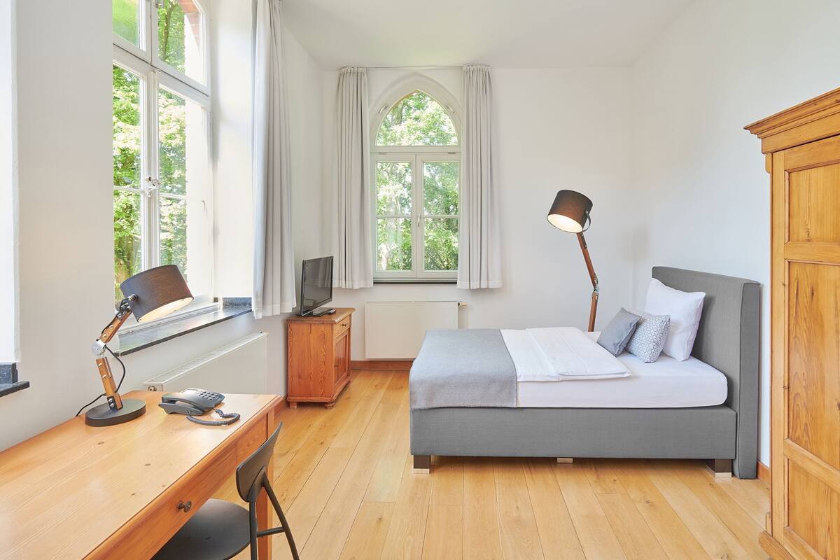 A bedroom with a bed, desk, chair, lamp, wardrobe, and large windows.