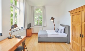 A bedroom with a bed, desk, chair, lamp, wardrobe, and large windows. - Hotel MutterHaus Düsseldorf (Düsseldorf)