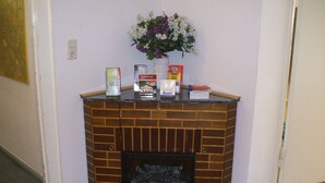 A corner fireplace with a brick design, a vase with flowers, and various books and a box on top. - Pension Messe am Funkturm (Berlin)
