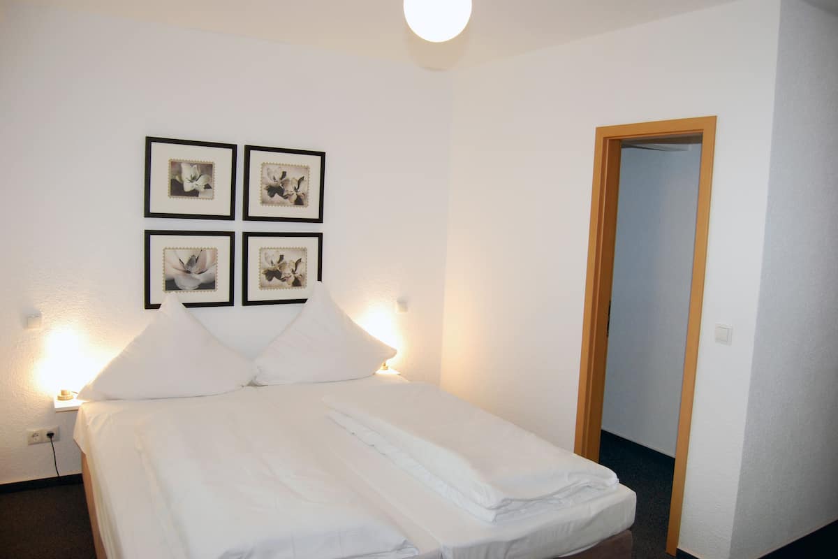 A neatly made bed with white linens, a wall-mounted light, and four framed pictures above it.