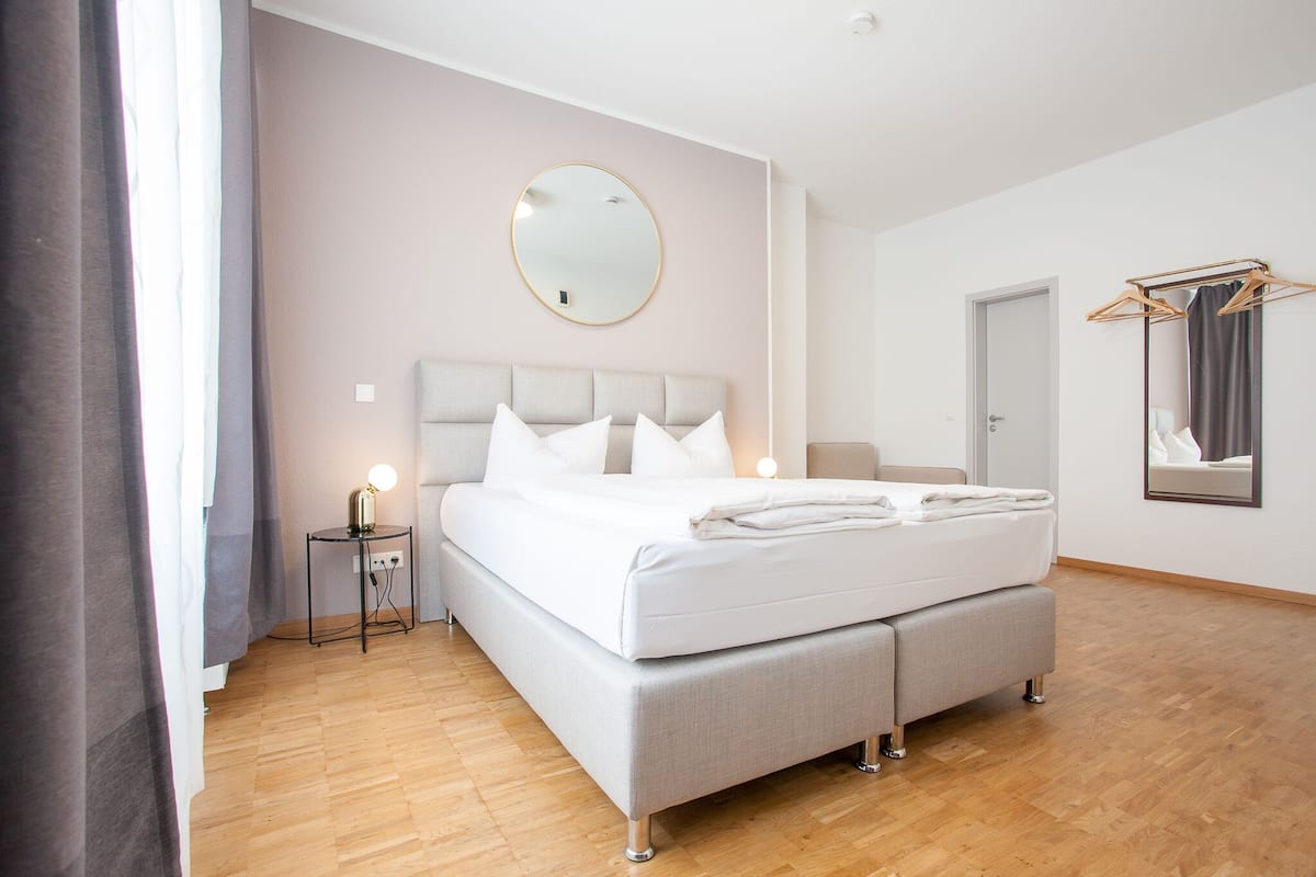 A modern bedroom with a large bed, bedside tables, a round mirror, and a wooden floor.