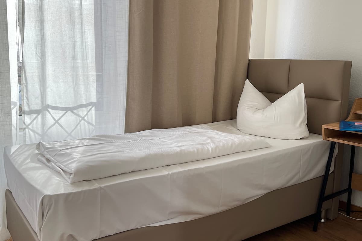 A neatly made bed with white linens in a room with wooden flooring and a window with sheer curtains.