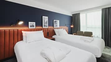 Hotellihuone, jossa on kaksi sänkyä, sängynpääty, ikkuna verhoilla ja seinään kiinnitetyt valaisimet.