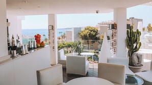 A modern outdoor seating area with white furniture, a cactus plant, and a view of the sea and buildings. - Marins Playa (Son Servera)
