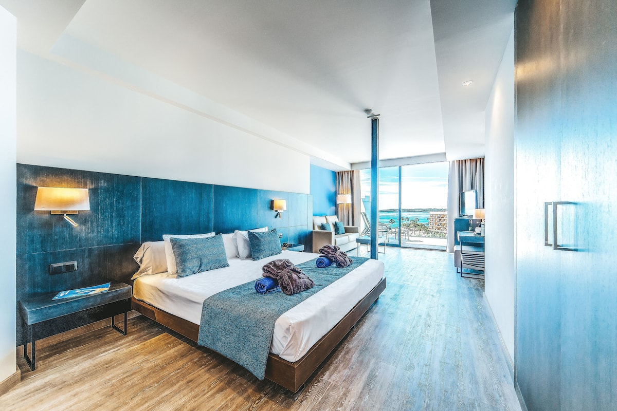 Habitación de hotel moderna con una cama grande, un banco y vistas al exterior.