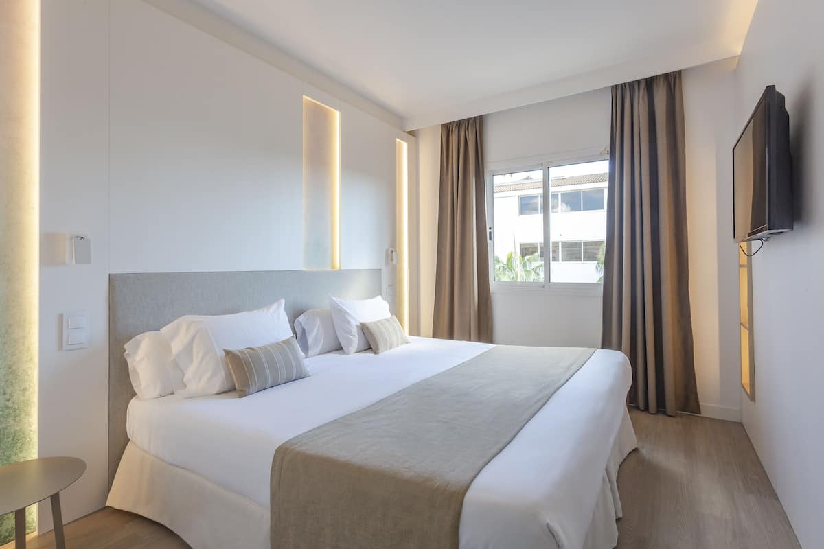 Una habitación de hotel moderna con una cama grande, un televisor de pantalla plana montado en la pared y un ventanal con cortinas.