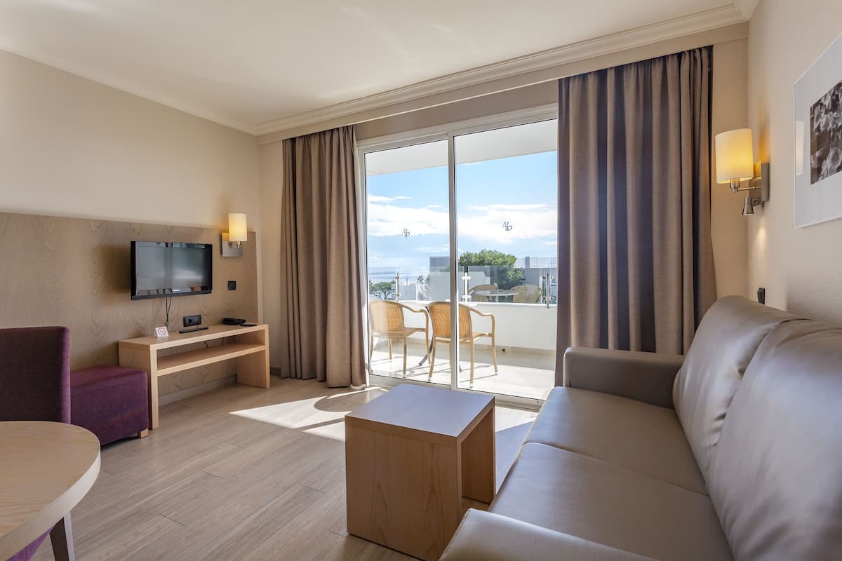 Una habitación de hotel moderna con sofá, una mesita de madera, televisor de pantalla plana y balcón con sillas.
