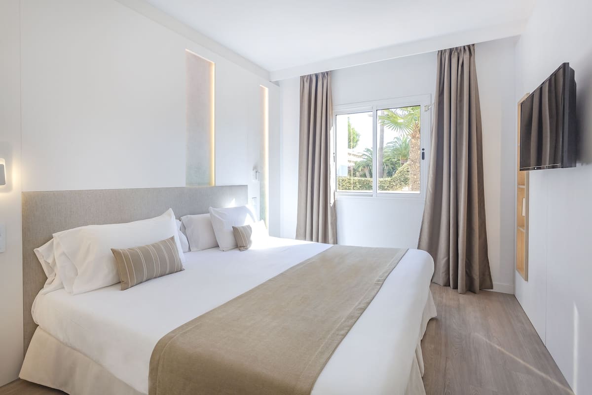 Una cama bien tendida con cabecera, un televisor montado en la pared y una ventana con cortinas.
