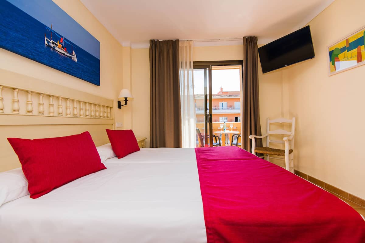 A hotel room with two beds, a TV, a balcony with a view of buildings, and a painting of a boat on the wall.