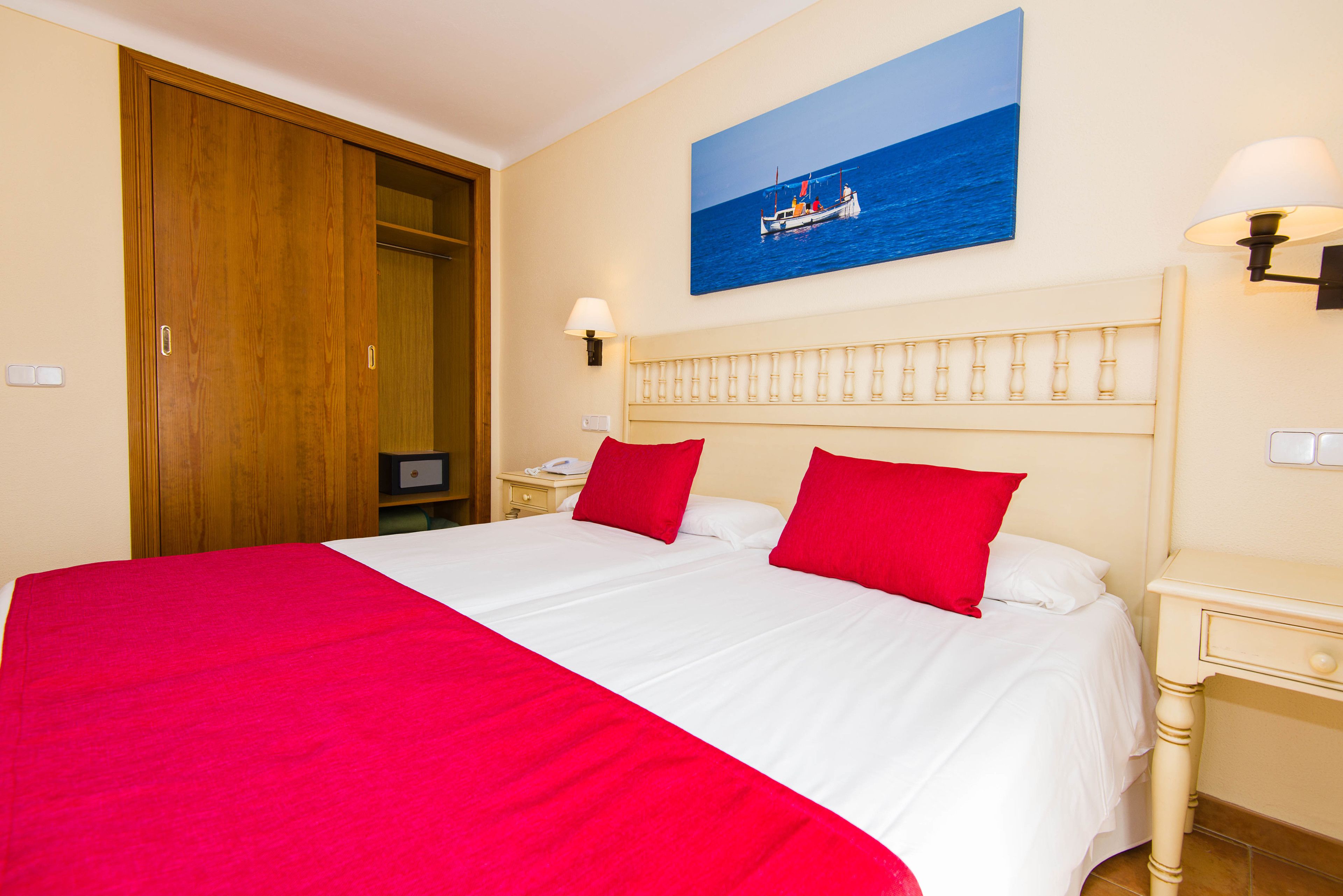 A hotel room with a large bed, red pillows, a wooden wardrobe, a nightstand with a lamp, and a painting of a boat on the wall.
