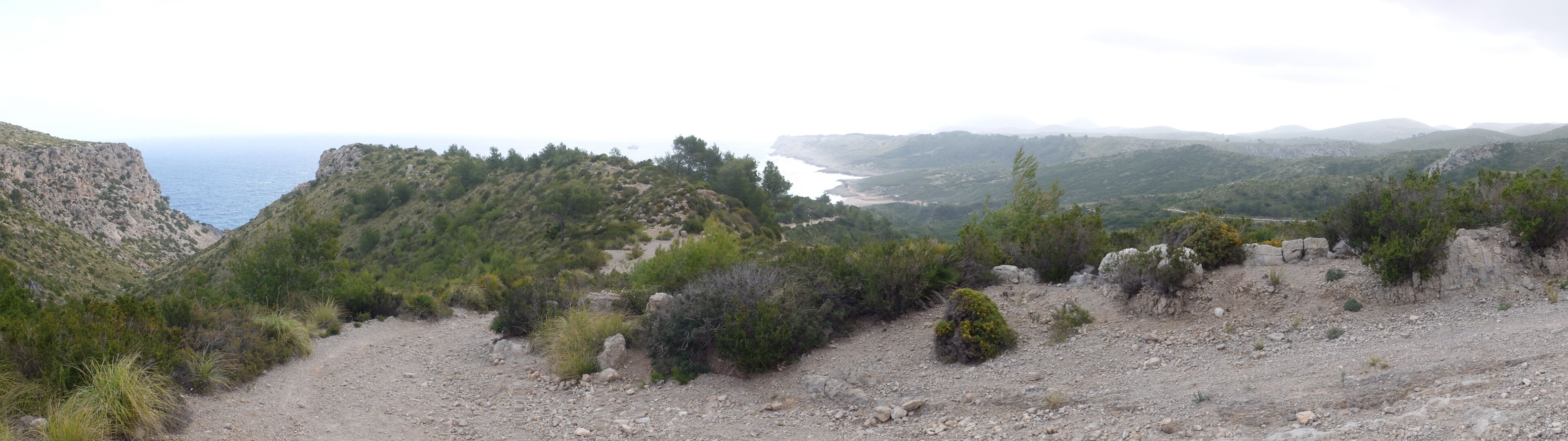 A rugged terrain with rocky paths and sparse vegetation, overlooking a coastal area with distant mountains and a body of water.