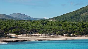 Eine Küstenlandschaft mit Strand, klarem blauen Wasser und einem bewaldeten Hügel im Hintergrund.