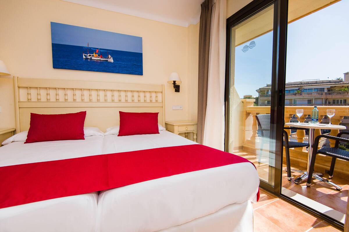 A hotel room with a large bed, red pillows, a painting of a boat on the sea, and a balcony with a table and chairs.