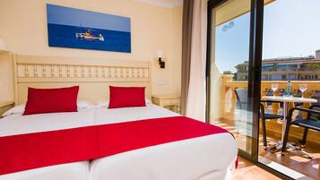Een hotelkamer met een groot bed, rode kussens, een schilderij van een boot op zee, en een balkon met een tafel en stoelen.
