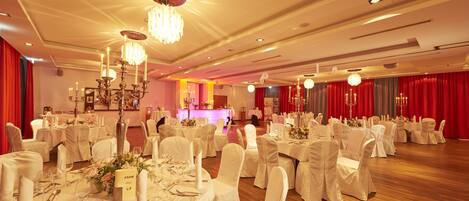 A banquet hall with round tables, white tablecloths, and chandeliers.