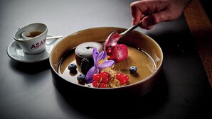 A dessert plate with a chocolate cake, berries, and a purple flower, accompanied by a cup of coffee labeled "ASAM".