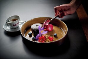 A dessert plate with a chocolate cake, berries, and a purple flower, accompanied by a cup of coffee labeled "ASAM".