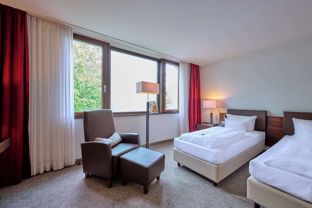 Quarto de hotel com duas camas, uma cadeira, uma escrivaninha e janela com cortinas.