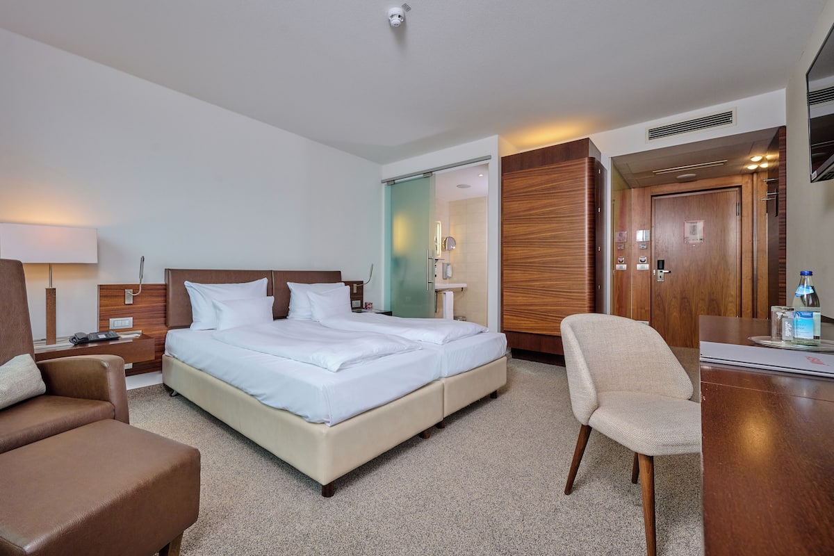 Quarto de hotel com uma cama grande, uma escrivaninha, uma cadeira e um banheiro visível através de uma porta de vidro.
