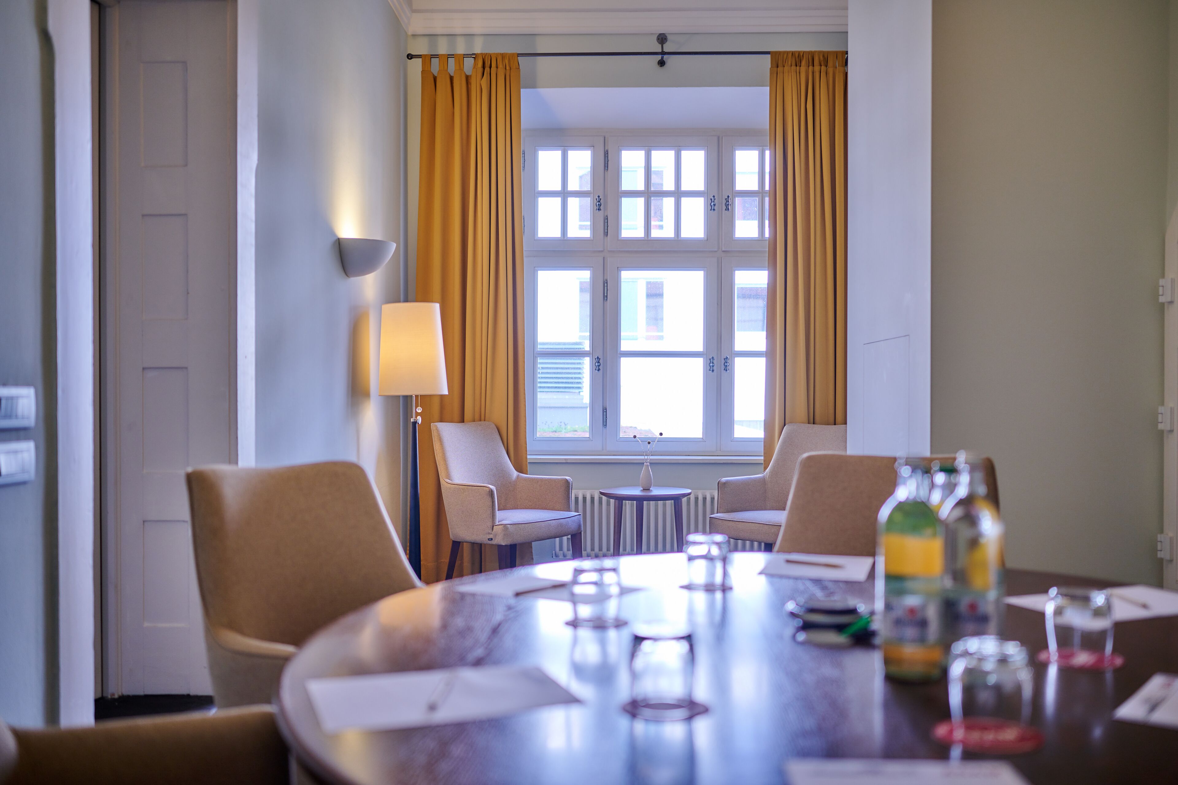 A conference room with a round table, four chairs, a lamp, a window with curtains, and a bottle of water on the table.